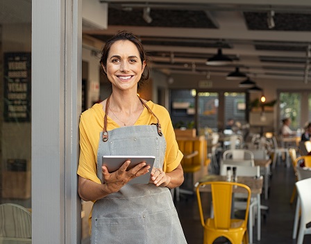 Portrait of happy woman standing at doorway of her store holding digital tablet. Cheerful mature waitress waiting for clients at coffee shop. Successful small business owner in casual clothing and grey apron standing at entrance and looking at camera.